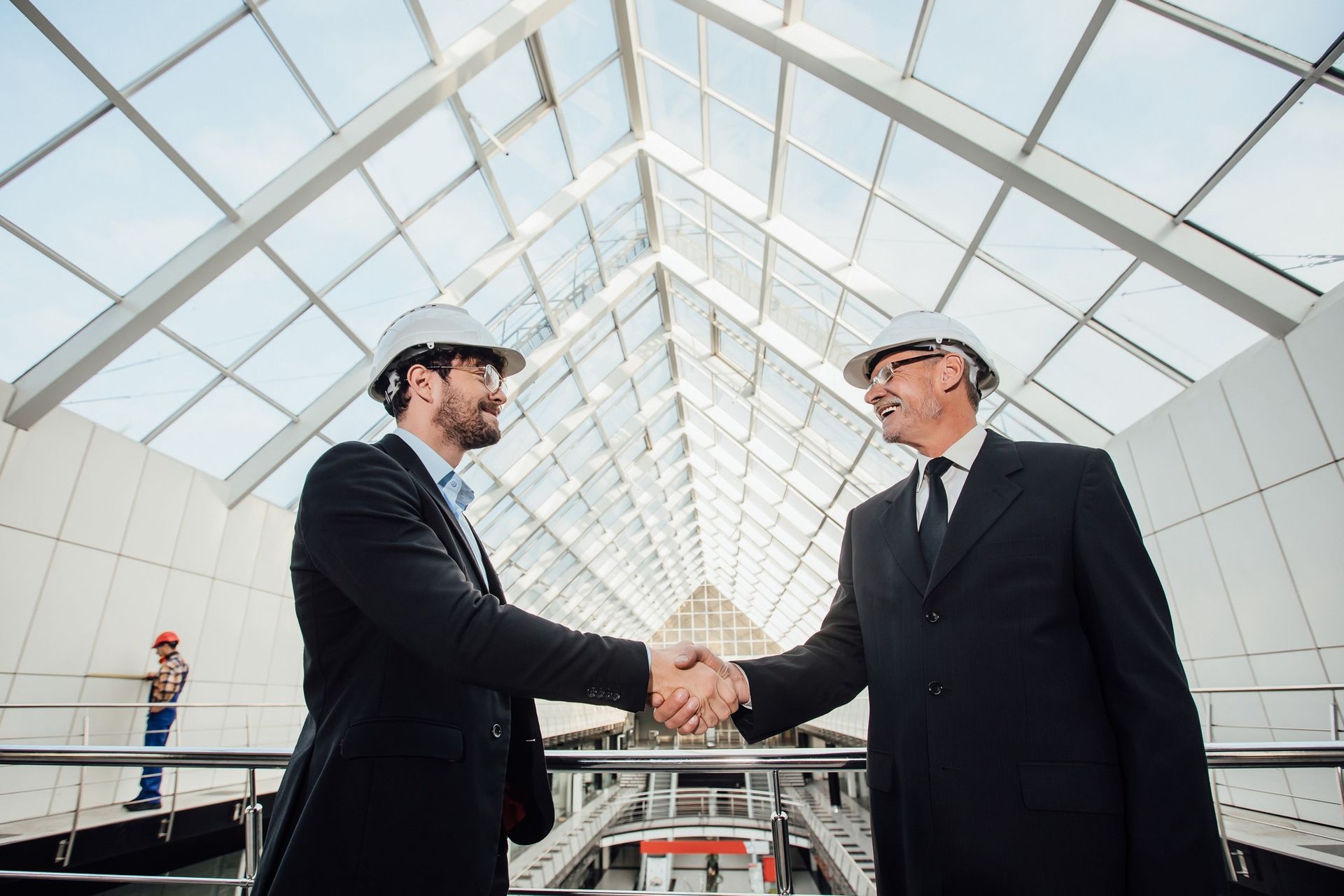 Two men in suits and hard hats shake hands under a glass roof, possibly after a deal.