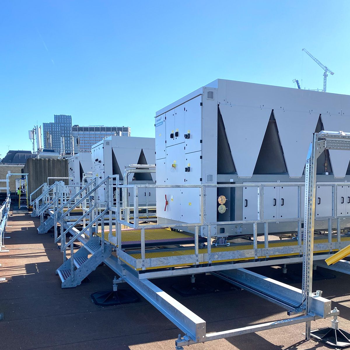 HVAC units on a rooftop platform, with stairs and railings for access. Clear blue sky, building in the background.