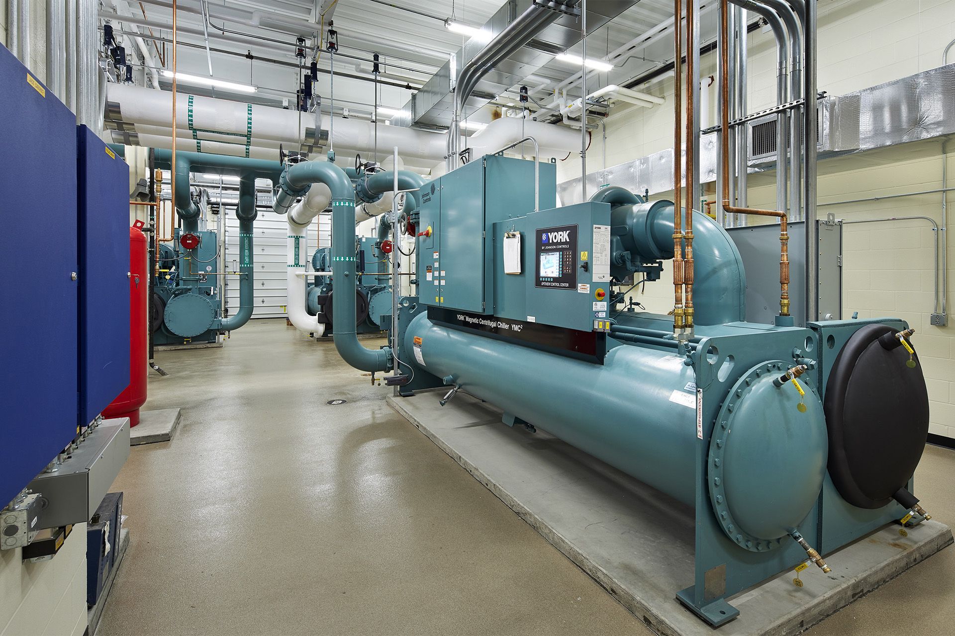 Equipment room with large teal chillers, pipes, and electrical panels.
