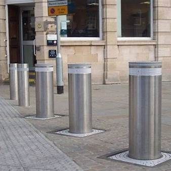 Five silver, retractable bollards on a sidewalk, likely for traffic control, near a building entrance.