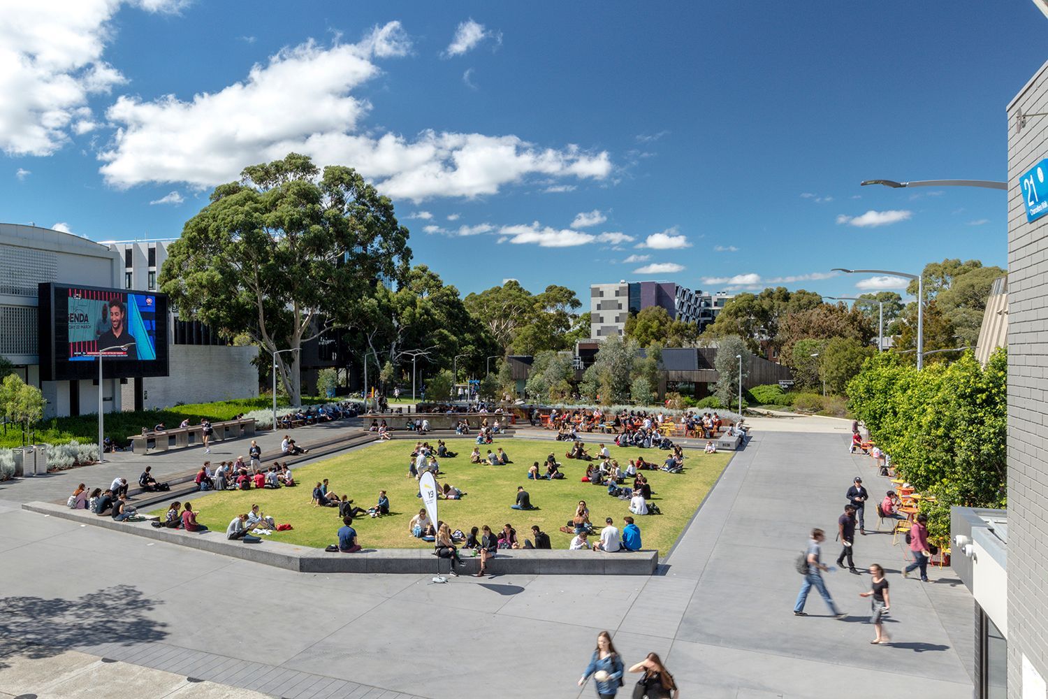People relax on the grass in a sunny campus courtyard. Buildings and blue sky with clouds are in the background.