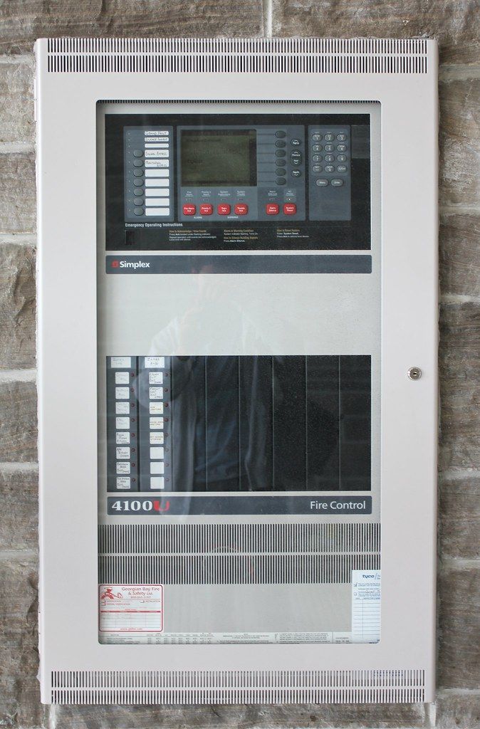 Fire control panel mounted on a brick wall, with a display screen, buttons, and other controls.