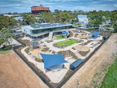 Aerial view of a modern playground with a blue canopy, landscaping, and a two-story building with solar panels.