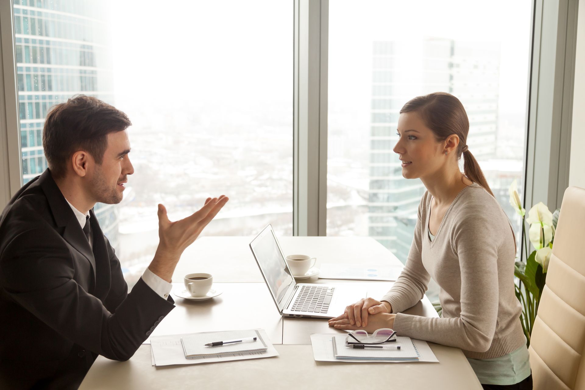 Man in suit gesturing, talking to woman in sweater at a table with laptop and coffee.
