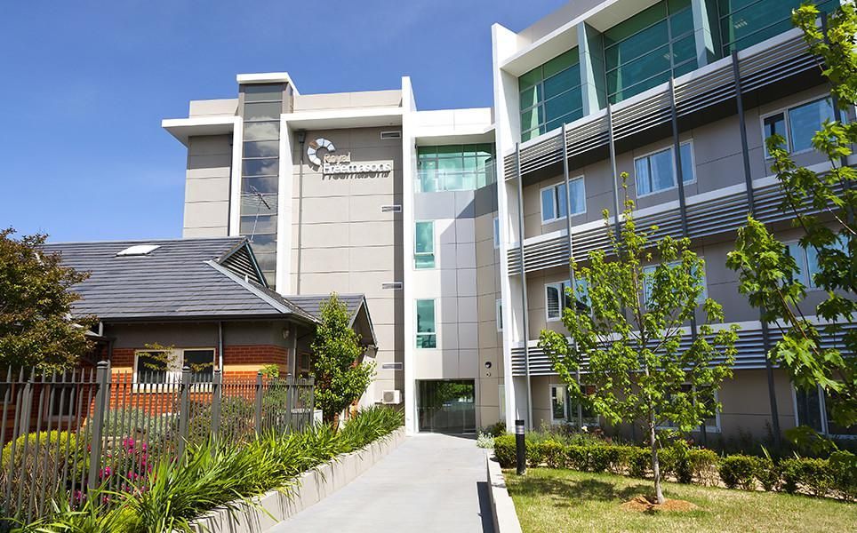 Modern building with glass windows and a walkway, next to a brick house and greenery.