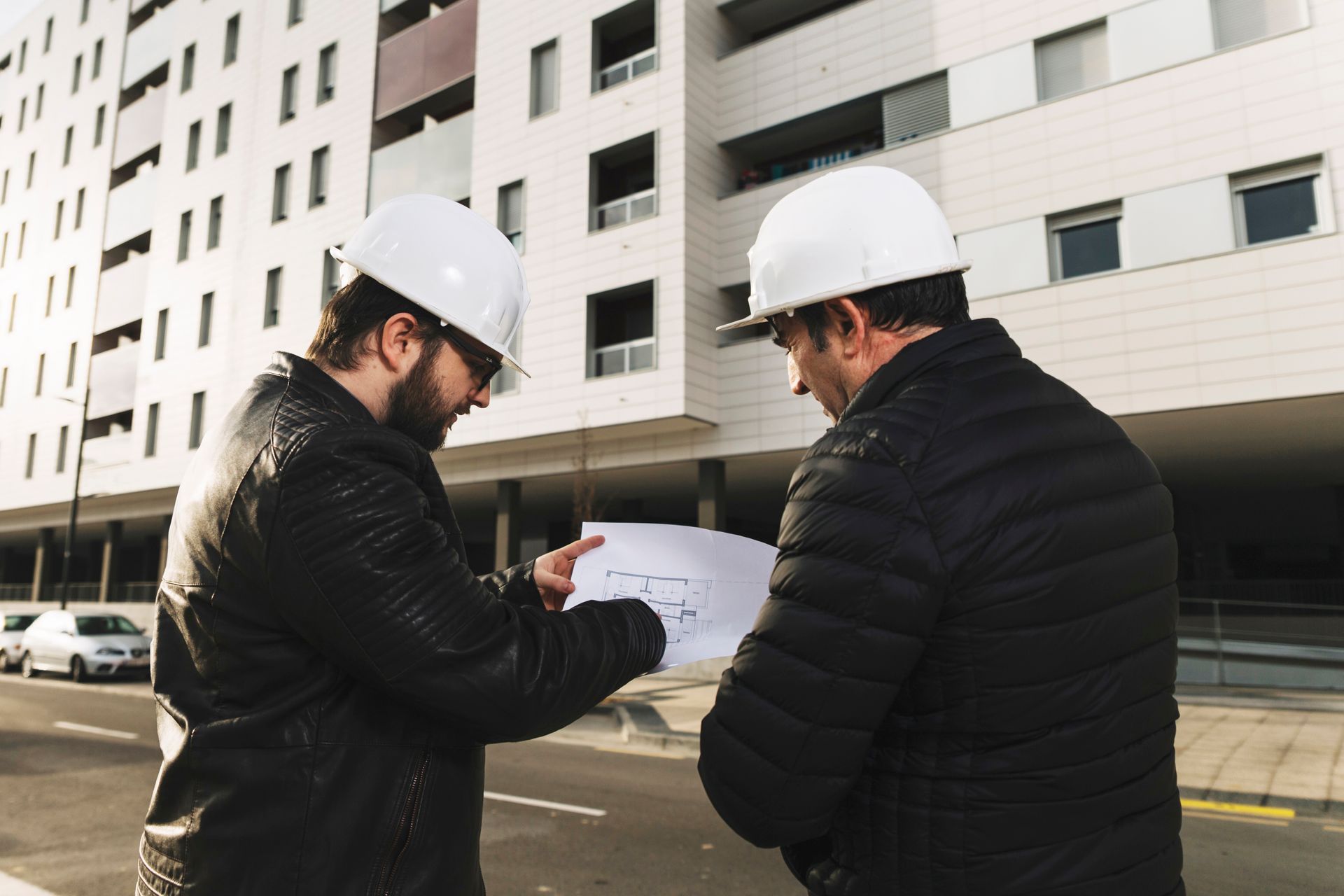 Two construction workers in hard hats examine blueprints in front of a building.