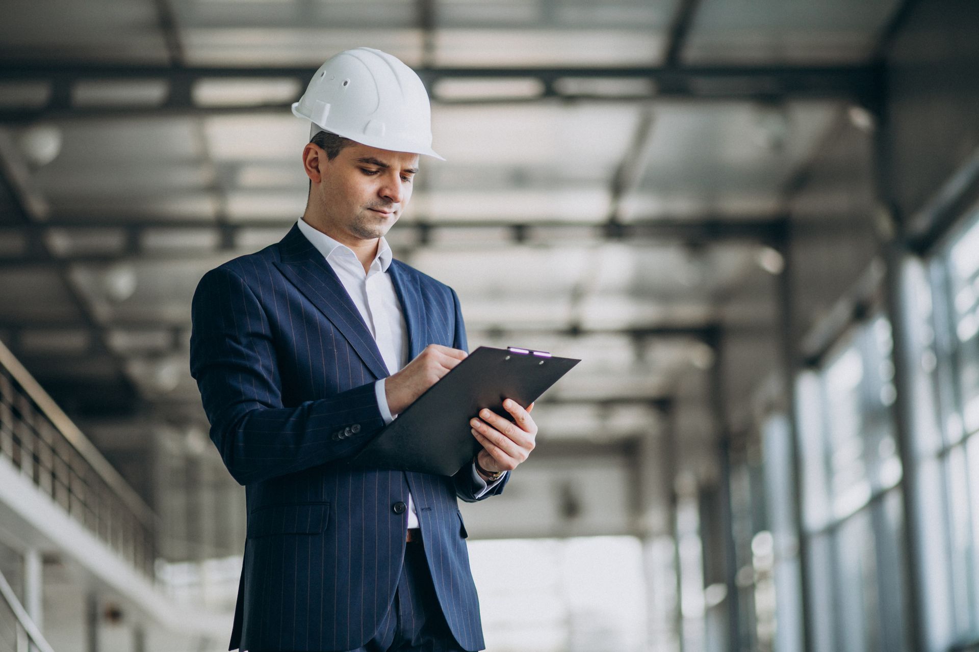 Man in suit and hard hat, writing on a clipboard in an industrial setting.