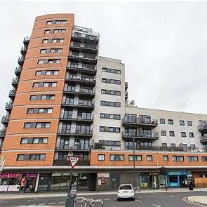 Multi-story brick and white apartment building with balconies and ground floor shops. A car is on the street.
