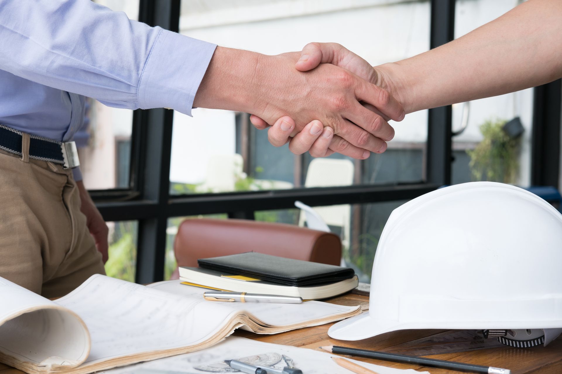 Two people shaking hands over a desk with blueprints, a hard hat, and a phone.