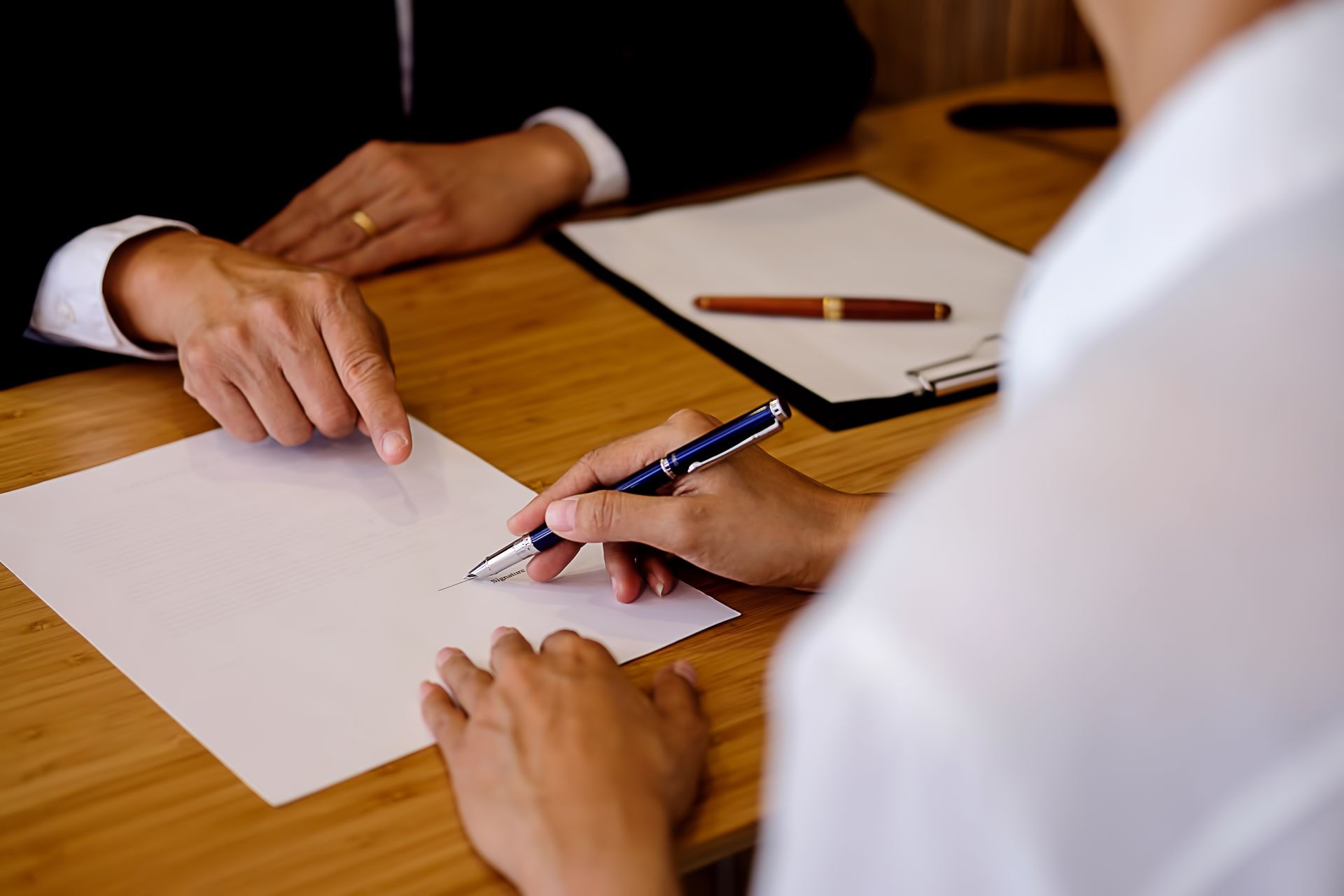 Two people at a table: one signing a document as another points, likely a contract signing.