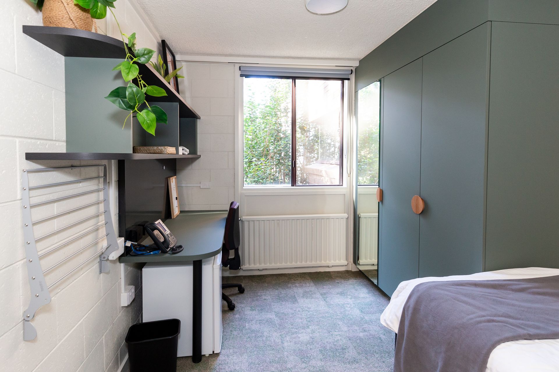 A bedroom with a desk, wardrobe, and window overlooking greenery.