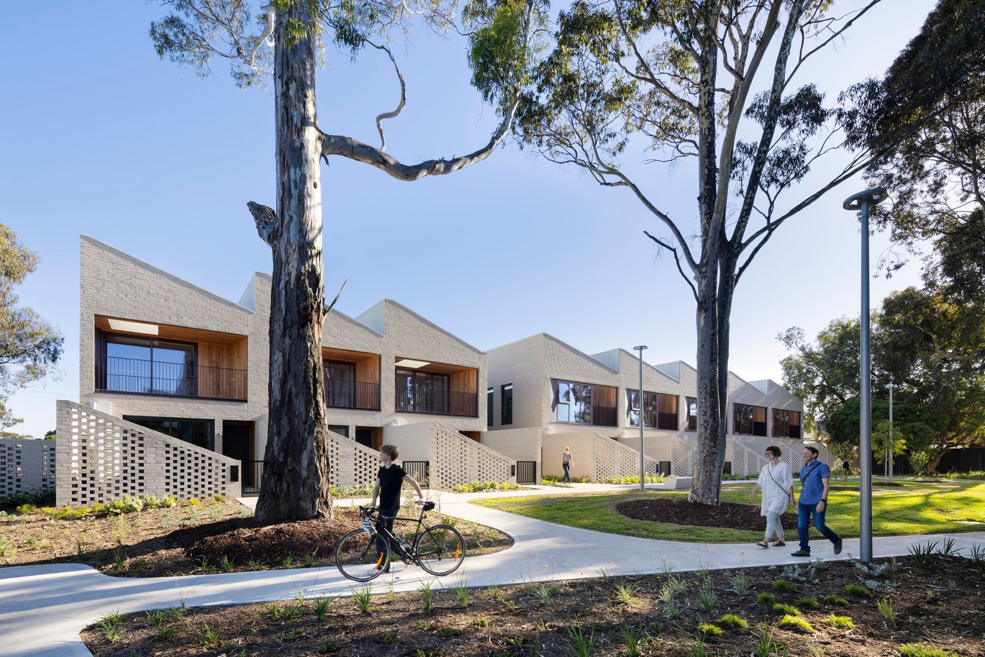 Row of light brick houses with angled roofs, trees, walkway. Two people walking. Sunny day.