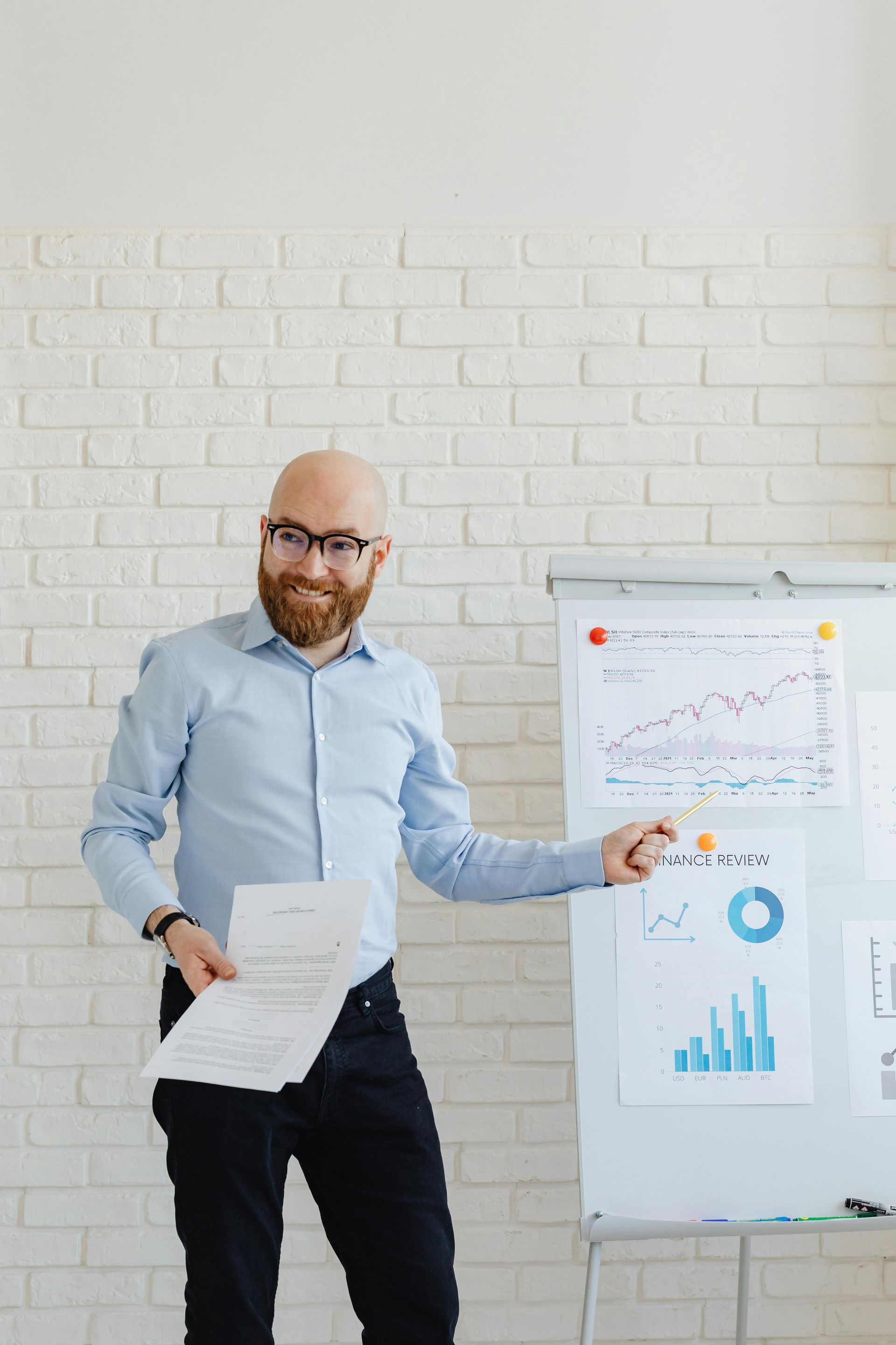 Bald man with a beard gestures at a whiteboard with charts while holding papers. White brick wall in the background.