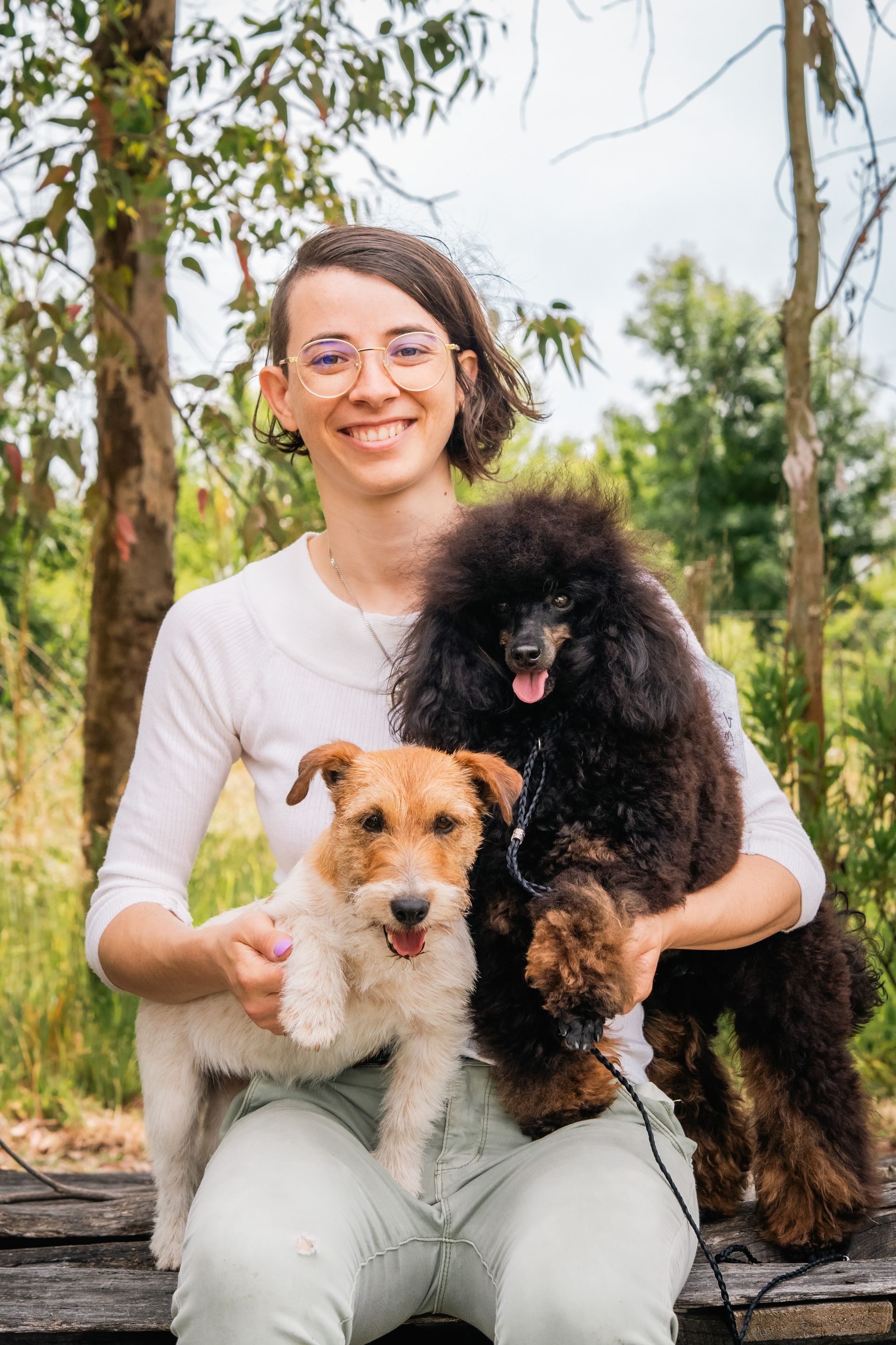 Donna sorridente con gli occhiali, che tiene in braccio un piccolo cane bianco e marrone chiaro e un grosso barboncino nero, all'aperto.