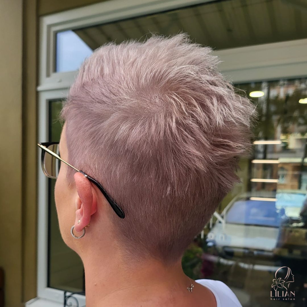 Eine Frau mit kurzen rosa Haaren und Brille steht vor einem Fenster.