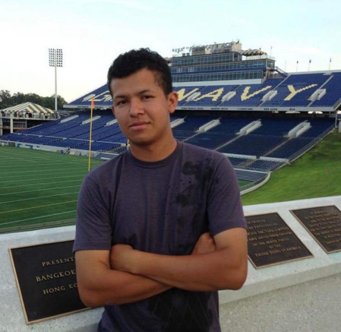 Man with arms crossed in front of a blue stadium, sunny day.