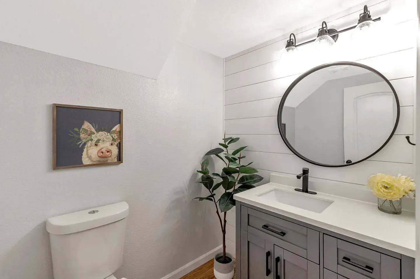 Bathroom with gray vanity, round mirror, pig art, and faux plant.