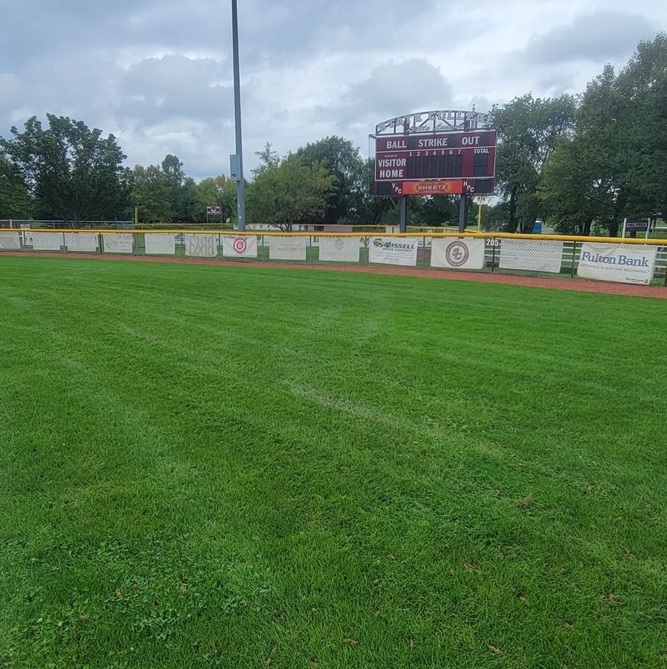 A baseball field featuring a lush green grass outfield, a dirt track, and a scoreboard under a cloudy sky.