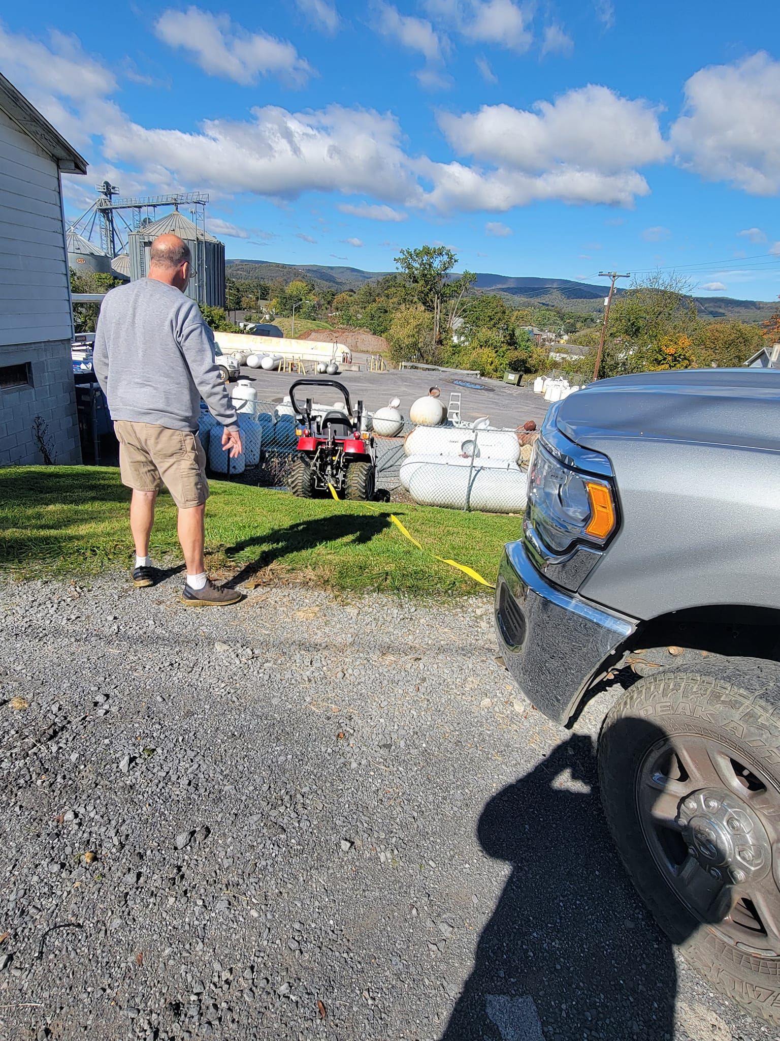 A person stands on a grassy lawn near a parked truck, looking toward a small red tractor and several large propane tanks.