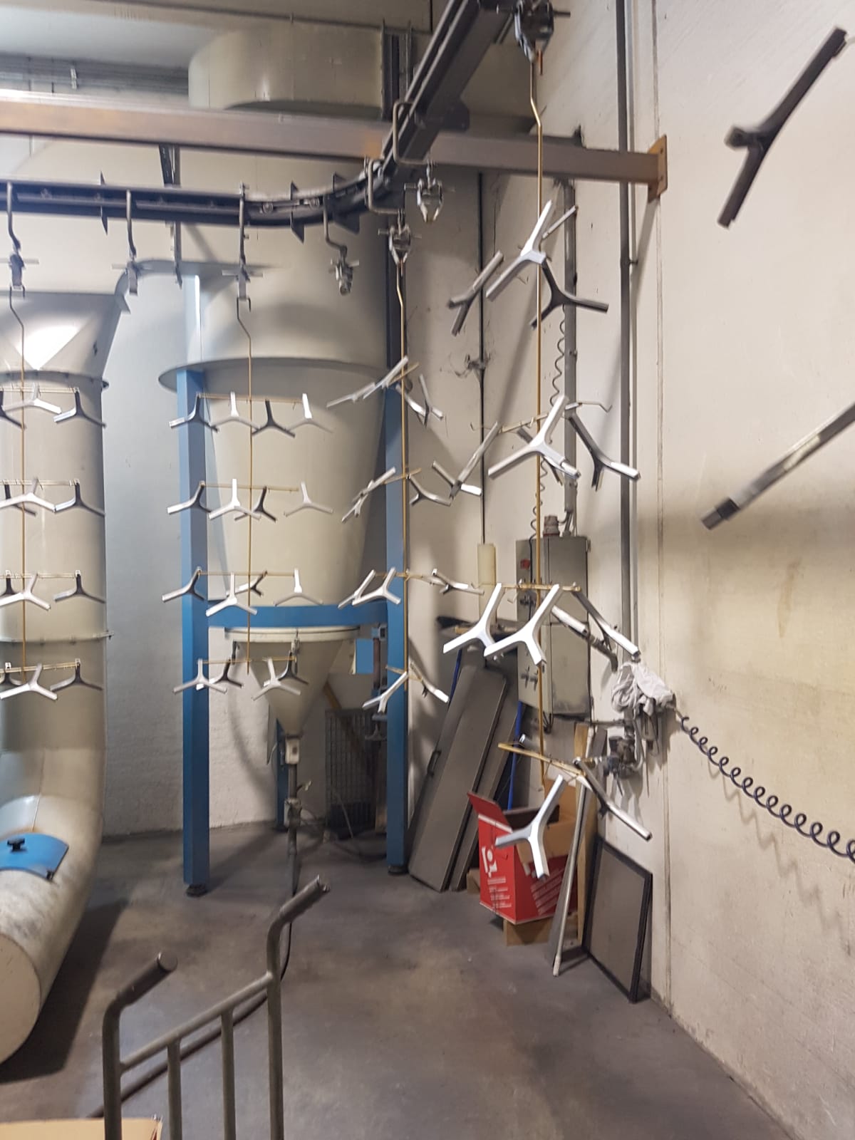 Locale tecnico industriale con tubature, telai di supporto blu, catene sospese e un secchio rosso vicino al muro.