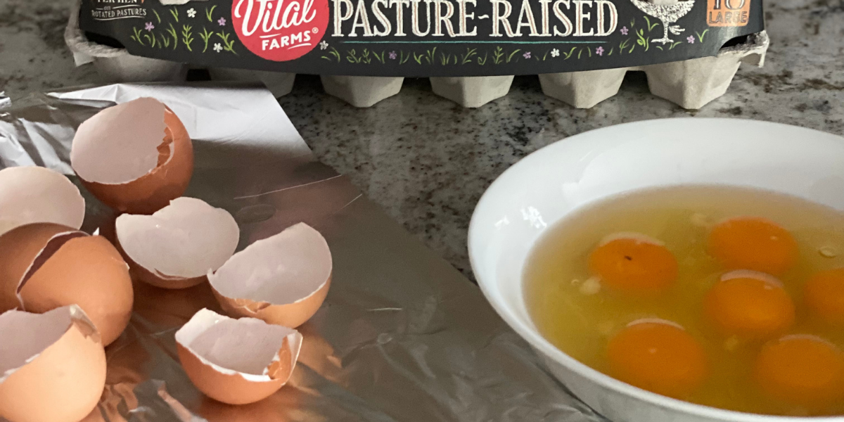 Egg shells and a bowl of cracked eggs are on a foil-covered surface. In the background is an egg carton.