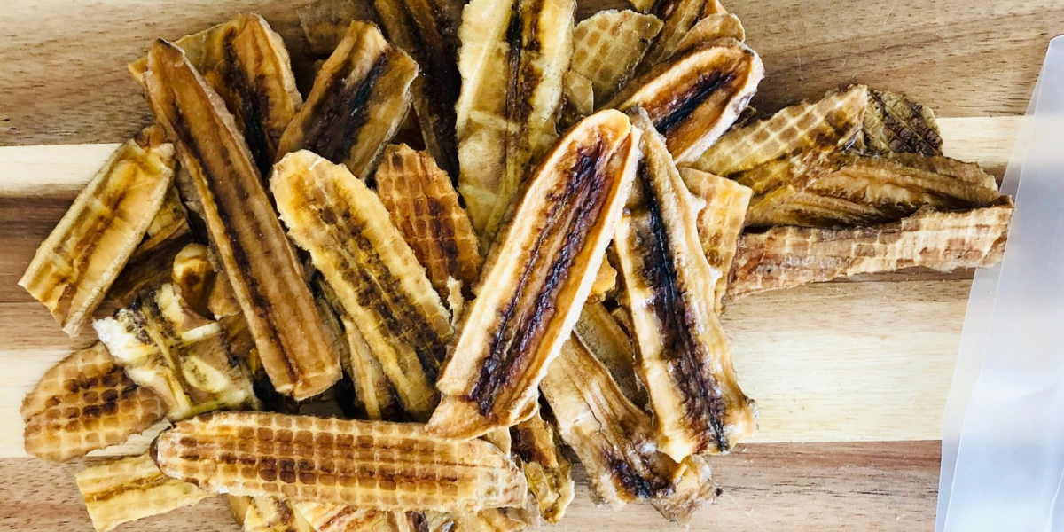 Pile of dehydrated banana slices on a wooden surface, with a blurred white object on the right side.