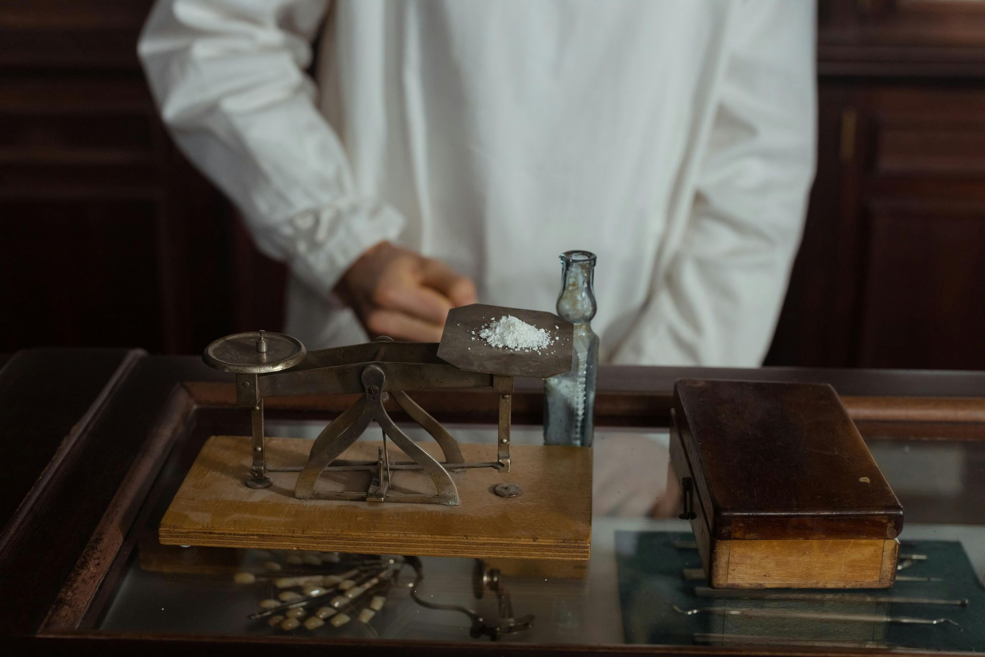 Person behind a counter weighing a white powder on a small scale, with a small bottle and wooden box nearby.