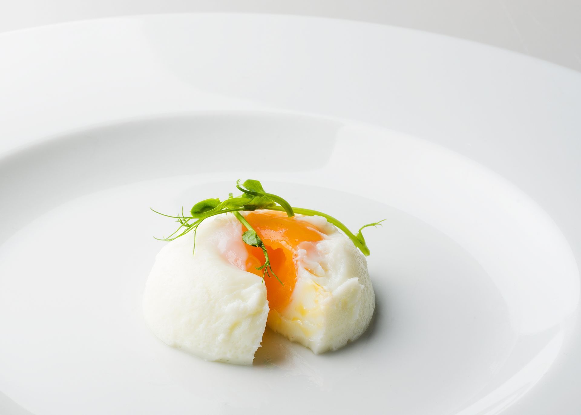 Poached egg with a bright orange yolk, garnished with green sprouts, on a white plate.