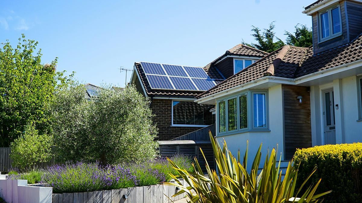 A house with solar panels on the roof is surrounded by trees and bushes.