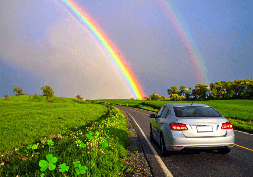 Sedan driving on a winding country road beneath a bright rainbow with shamrock graphics in the grass