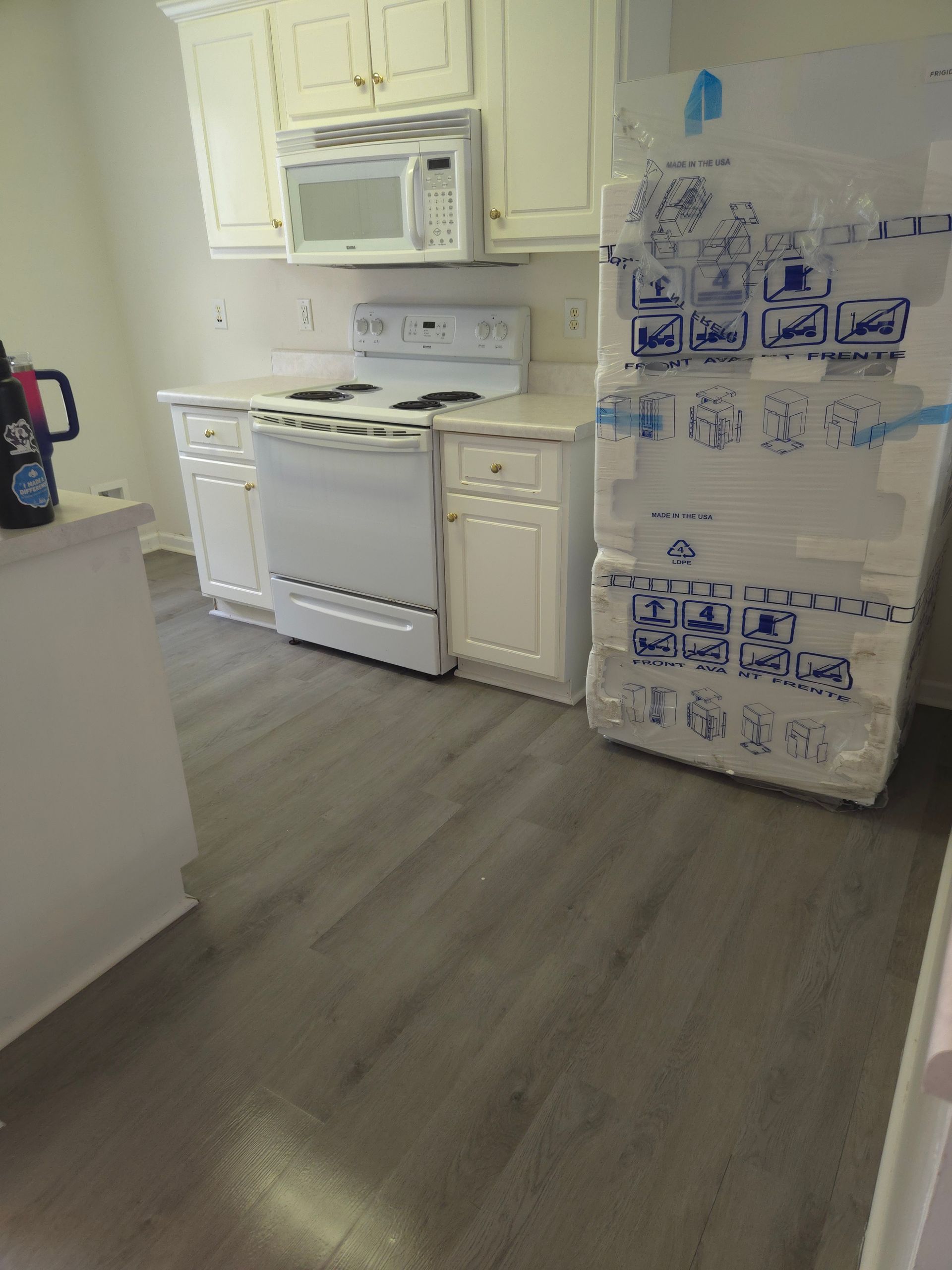 White kitchen with cabinets, stove, microwave, and new gray flooring. A new refrigerator is wrapped in protective packaging.