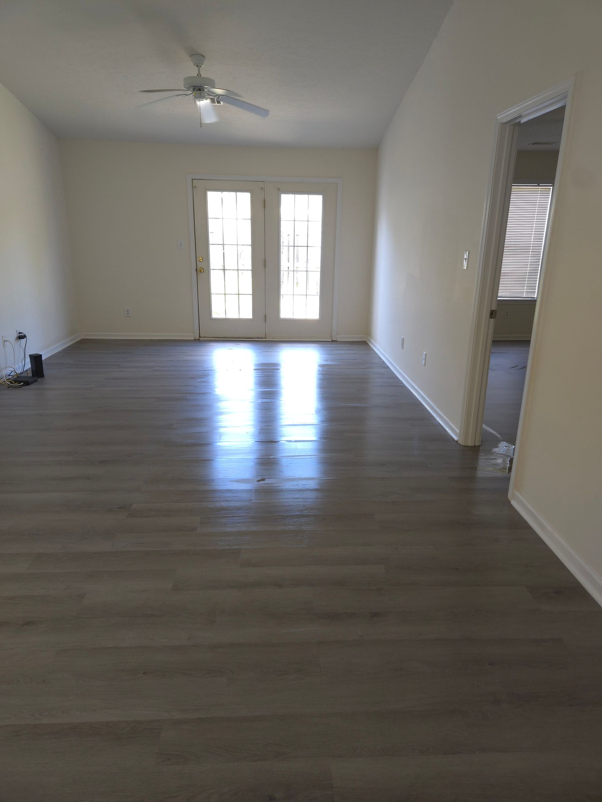 A bright living room with gray wood-look flooring, white walls, and French doors. Sunlight reflects off the floor's surface.