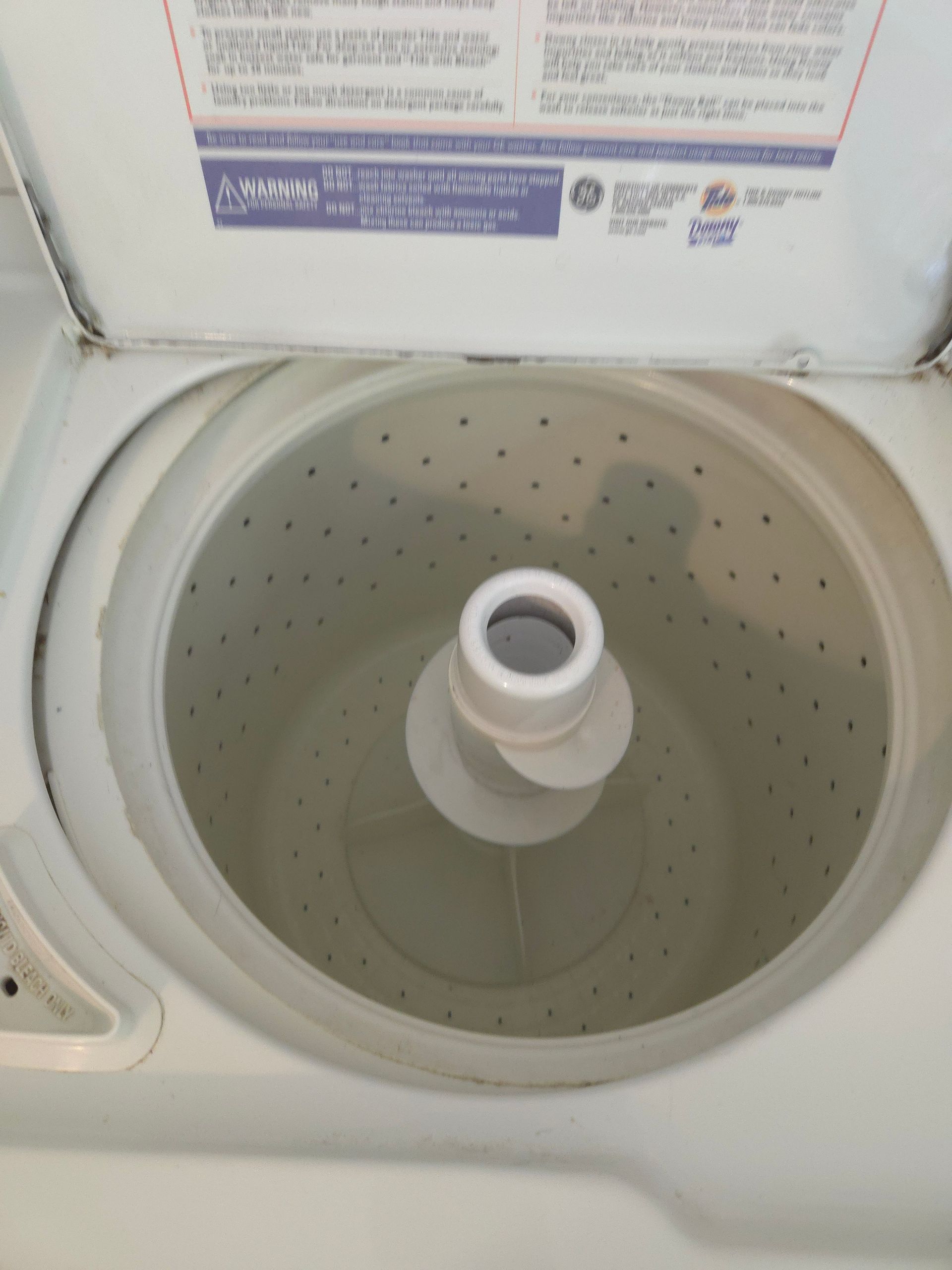 An open, white top-load washing. The central inner drum is visible, with a partially open lid in the background.
