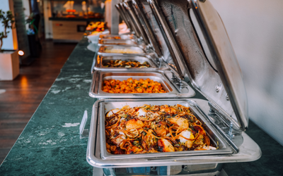 Buffet line with stainless steel trays of hot food, lids open on a counter in a restaurant