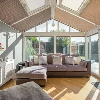 Living room with large windows and brown shades, light brown wood floor, and a brown sofa with gray pillows.