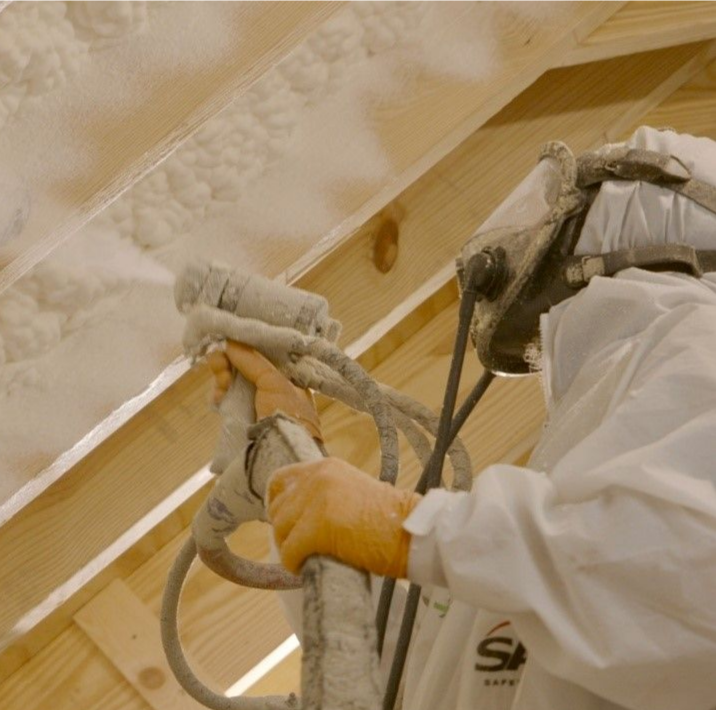 Person spraying insulation foam in an attic, wearing protective gear.