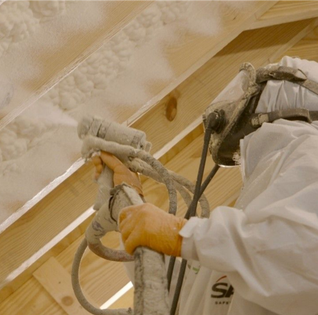 Person spraying insulation foam in an attic, wearing protective gear.