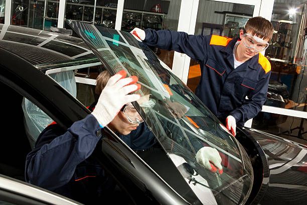 Two auto service technicians carefully lowering a new glass windshield onto a black vehicle.