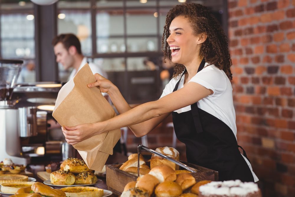 Woman in apron laughing, holding a paper bag, near baked goods at a cafe counter.