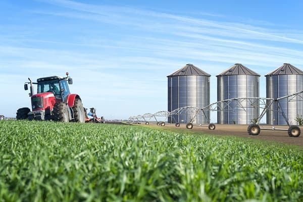 Um trator vermelho está passando por um campo com silos ao fundo.