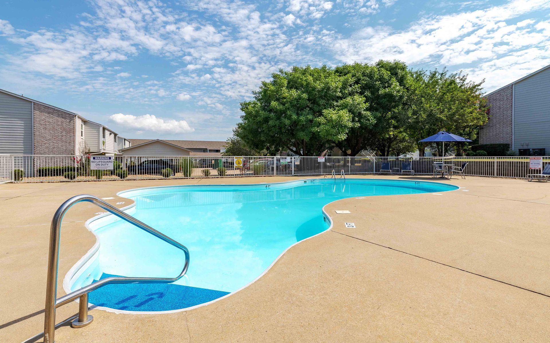 Swimming pool in an apartment complex on a sunny day. Blue water, beige deck, sky with clouds.