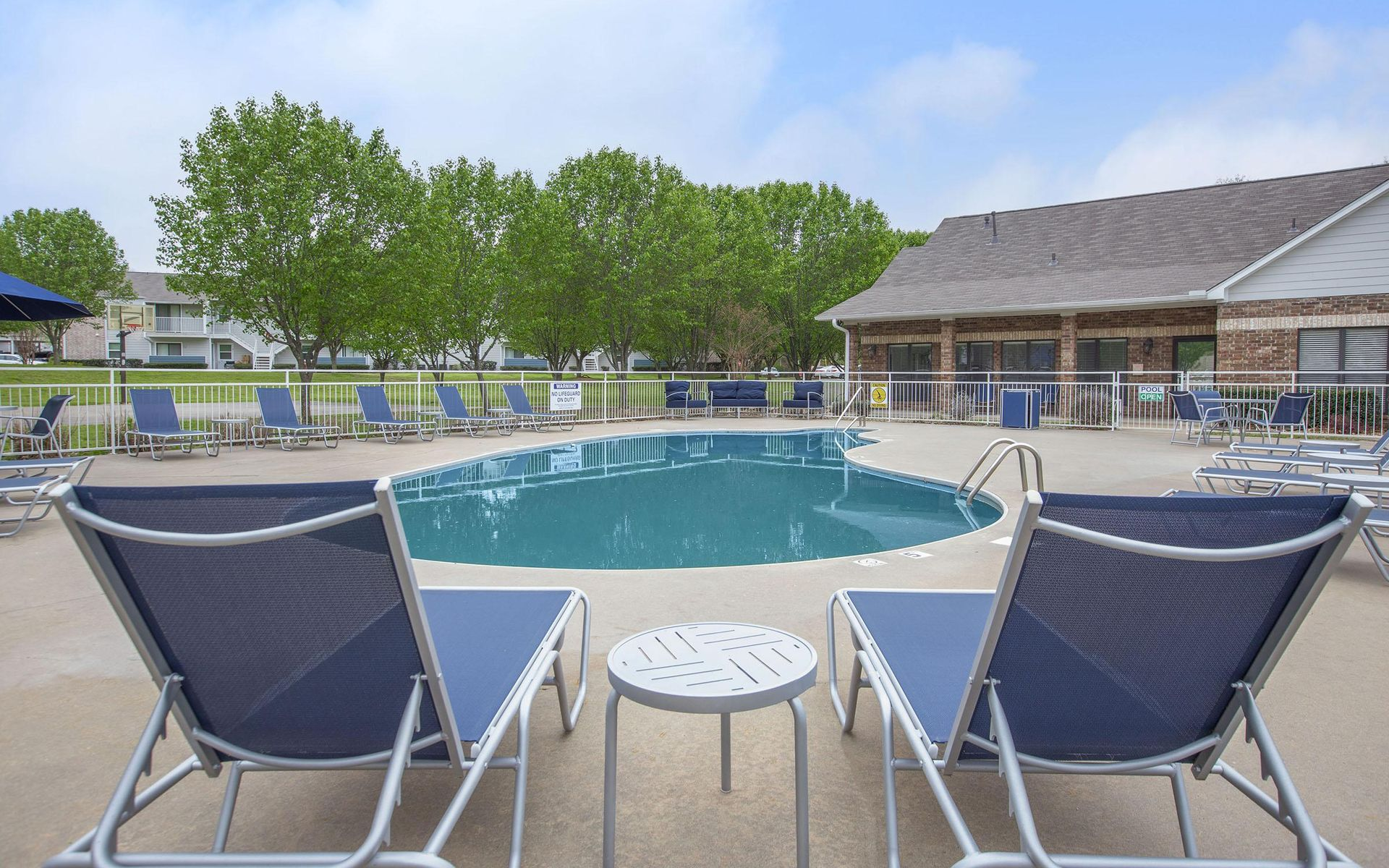 Two blue lounge chairs face a pool with a small table between them; covered patio and trees in background.