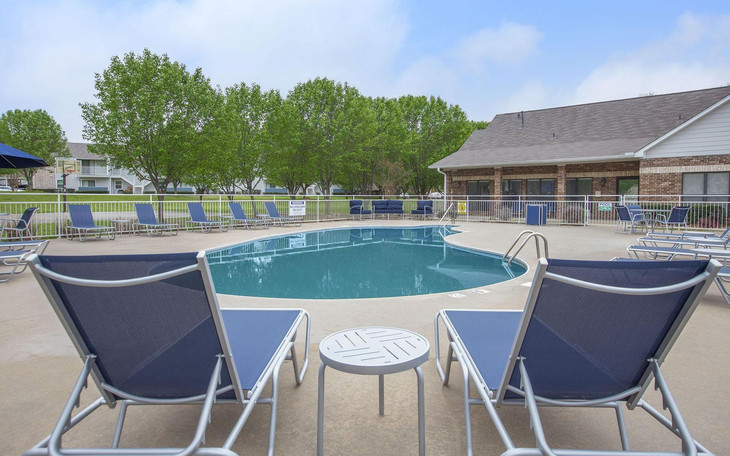 Two blue lounge chairs face a pool with a small table between them; covered patio and trees in background.