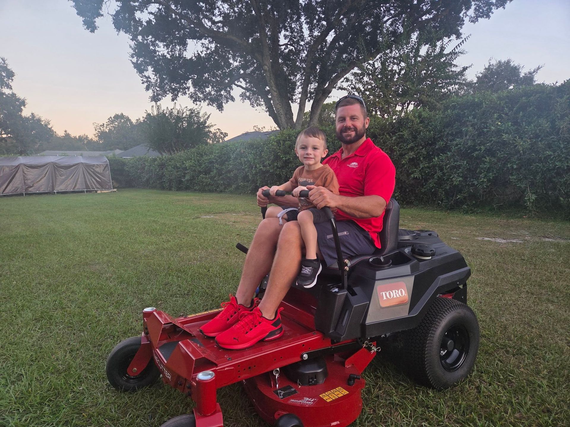 Man and child on a red riding lawn mower in a backyard, smiling.