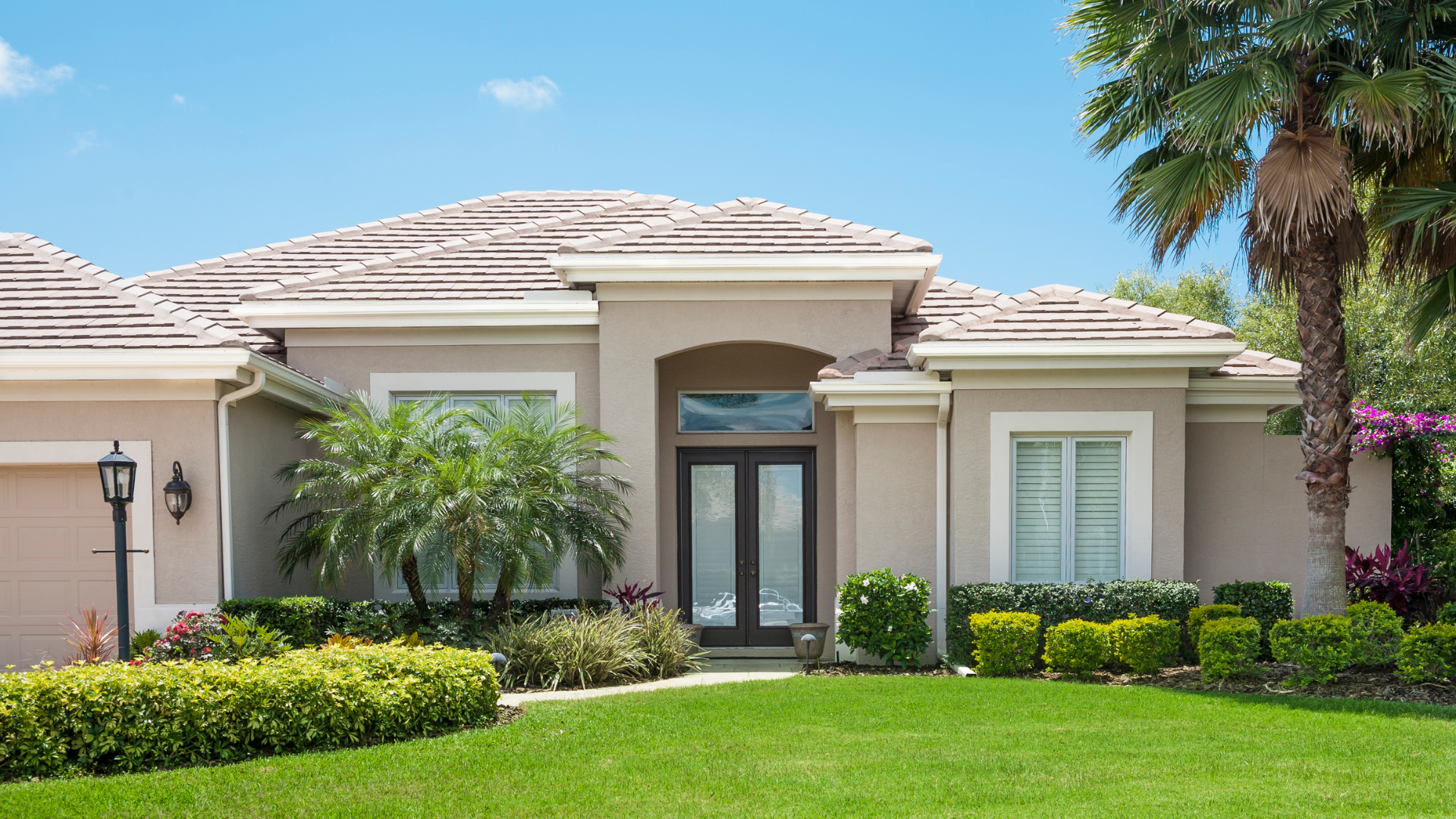 Beige stucco house with tile roof, green lawn, landscaping, and clear blue sky.