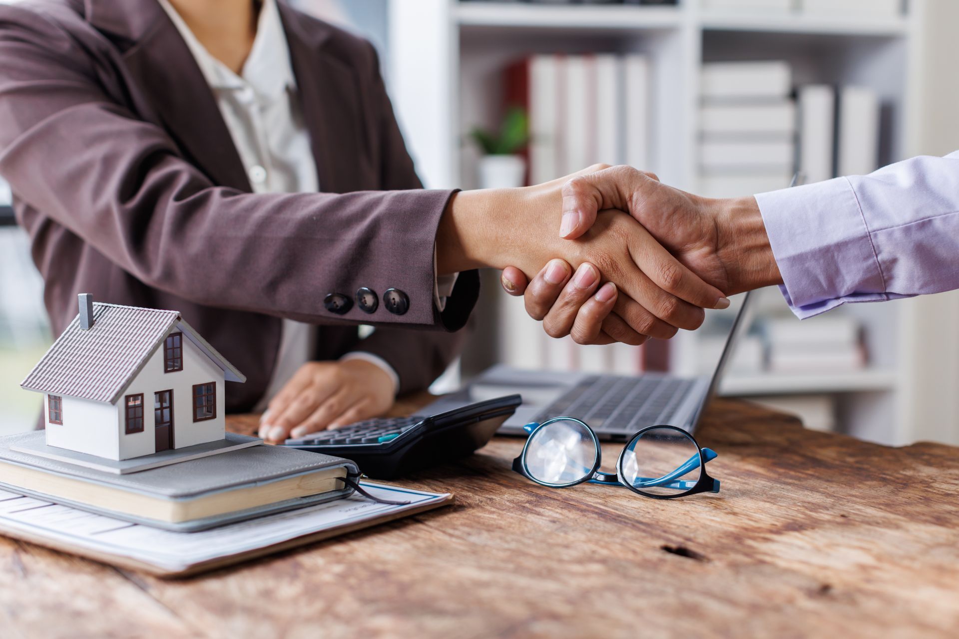 Two people shaking hands over a table with a small house model, keys, and contract papers