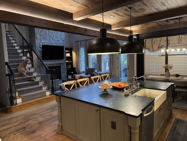Kitchen with island, dining area, and living room visible. Dog on stairs. Wooden beams and pendant lights.