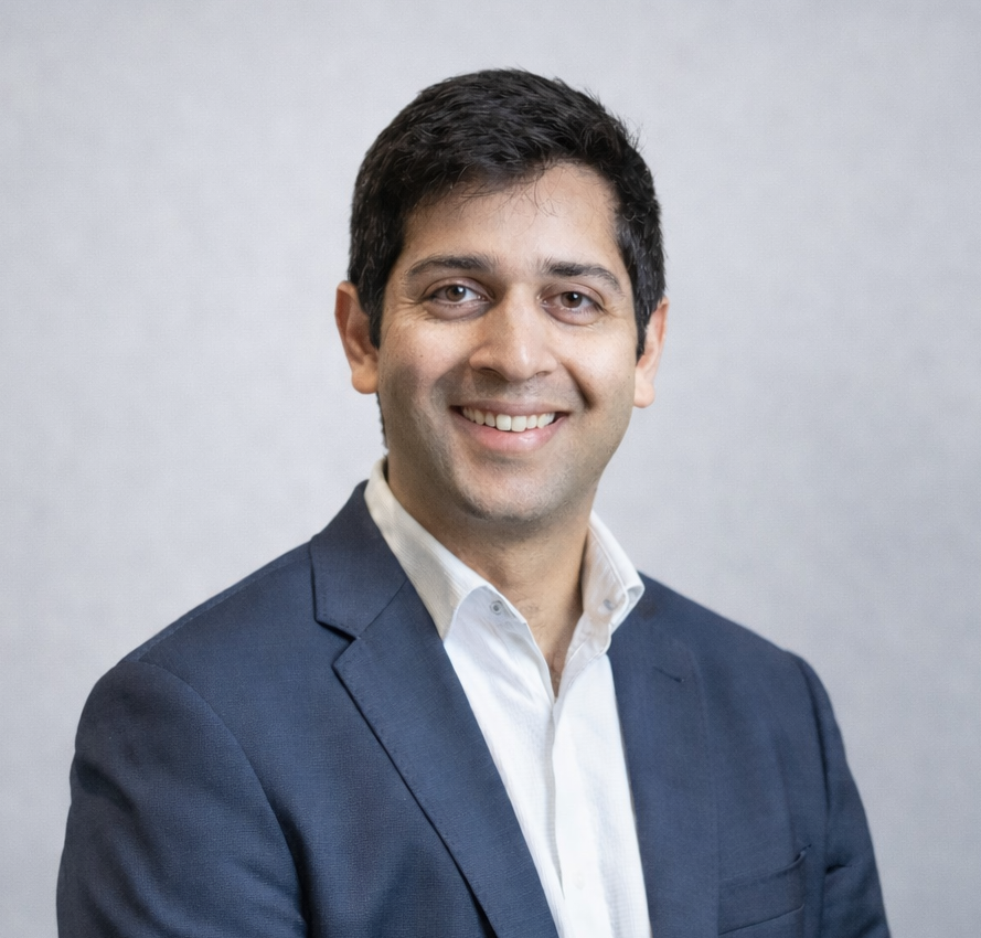 A man in a suit and white shirt is smiling in front of a wooden wall.