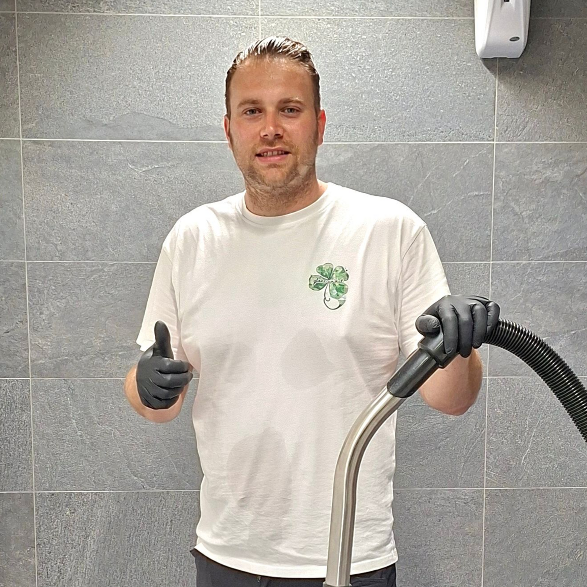 Mann im weißen Hemd mit Kleeblatt-Logo, der einen Staubsauger hält, zeigt den Daumen nach oben. Grau geflieste Wand.