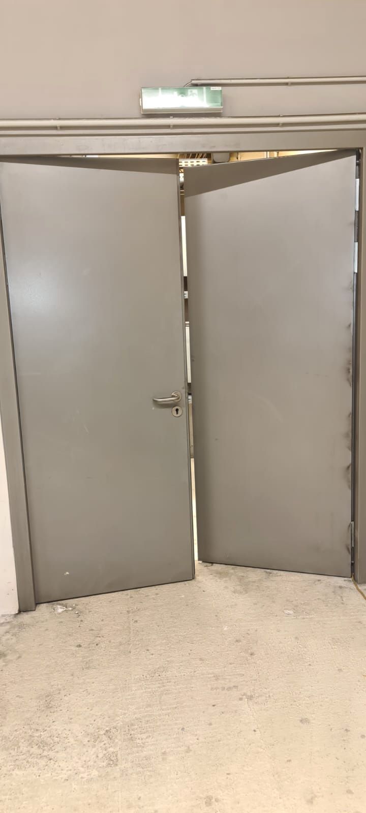 Zwei graue Türen, leicht geöffnet, in einem neutral gestrichenen Flur. Über den Türen ist ein Notausgangsschild zu sehen.